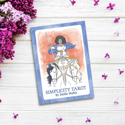 A tarot card deck titled "Simplicity Tarot" by Emilie Muñiz lies on a white wooden surface. The cover features an illustration of a woman with a spear riding a white and black horse, reminiscent of the Rider-Waite tradition. Purple and white flowers and small petals surround the Simplicity Tarot.