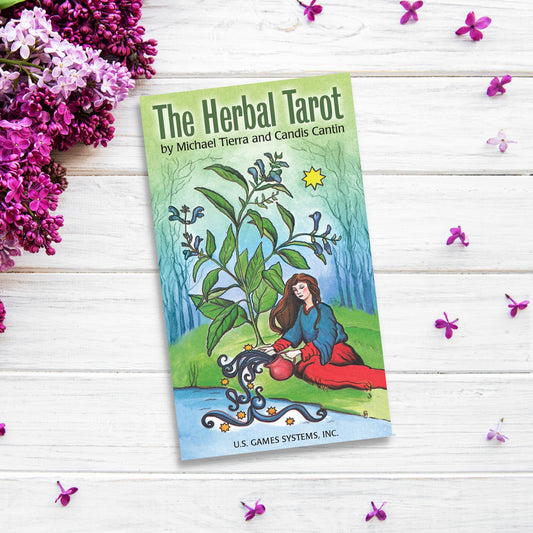 A colorful tarot card deck titled "The Herbal Tarot" by Michael Tierra and Candis Cantin is placed on a white wooden surface surrounded by vibrant purple flowers. The cover, enriched with medicinal herbs, features an illustration of a woman watering a large plant.
