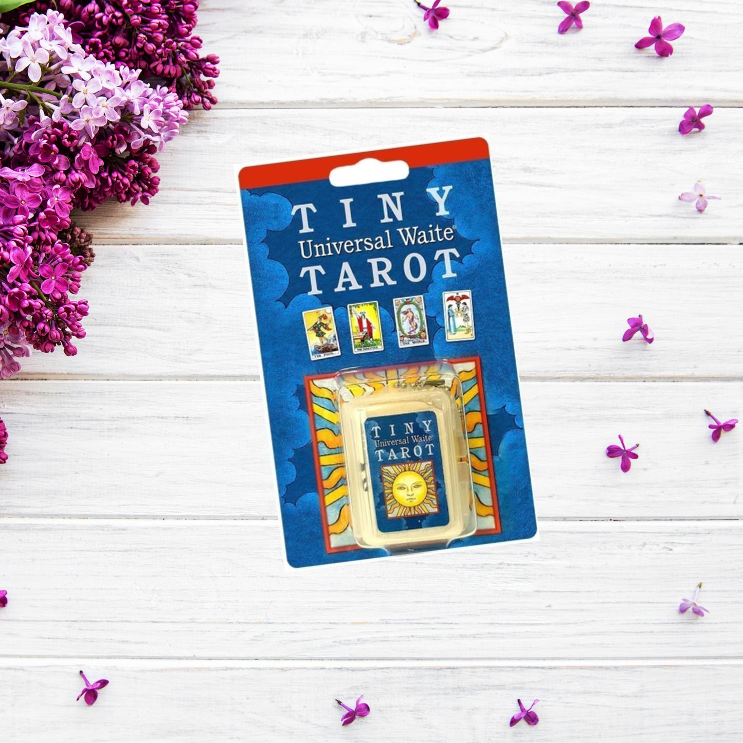 A pack of "Tiny Universal Waite Tarot Keychain" cards is displayed on a white wooden surface. The blue packaging features illustrations of four tarot cards and an image of a sun. Purple lilac flowers and scattered petals decorate the background, enhancing the beauty of this miniature tarot deck inspired by Pamela Colman Smith's work.