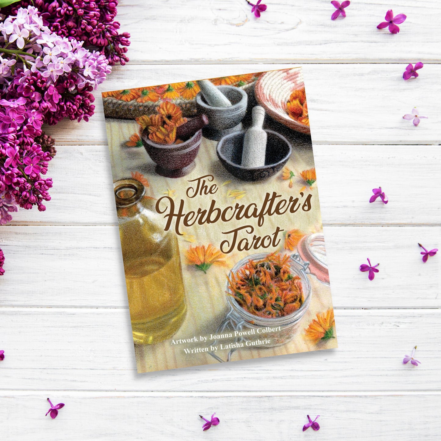 A book titled "The Herbcrafter’s Tarot" sits on a white wooden surface, surrounded by scattered purple flowers and a bunch of lilacs. The cover features bowls, herbs, and a mortar and pestle, emphasizing its focus on herbalism. Created by Joanna Powell Colbert and Latisha Guthrie.