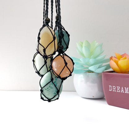 A collection of five polished stones of various colors, each encased in a handwoven black cord, hanging side by side. In the background, two small potted succulents with blue-green and orange leaves rest on a white surface. The product is called Empty Net Cage Necklace.