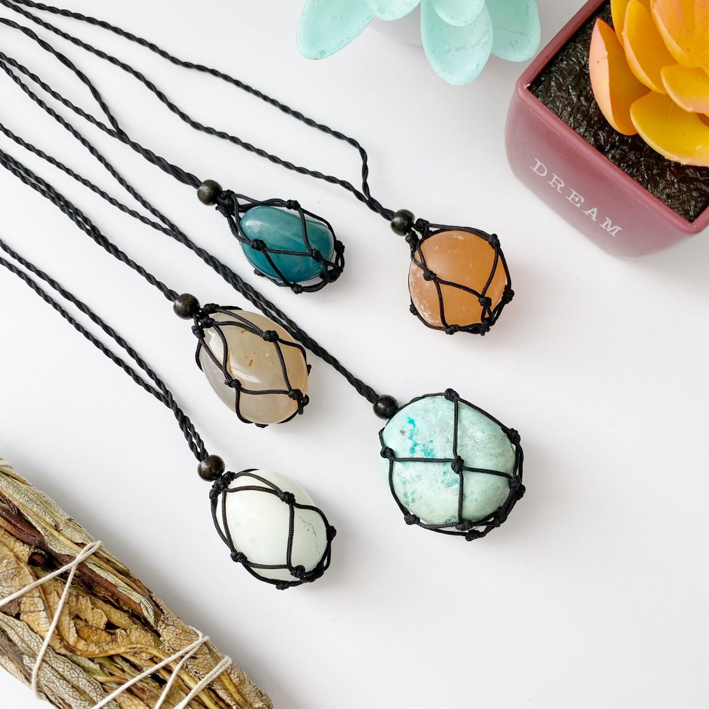 A collection of five polished stones of various colors, each encased in a handwoven black cord, hanging side by side. In the background, two small potted succulents with blue-green and orange leaves rest on a white surface. The product is called Empty Net Cage Necklace.