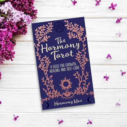 A tarot deck box titled "The Harmony Tarot" by Harmony Nice is placed on a white wooden surface. Surrounding the box are small purple flowers and some lilac blossoms, enhancing the serene and mystical vibe, perfect for personal growth.