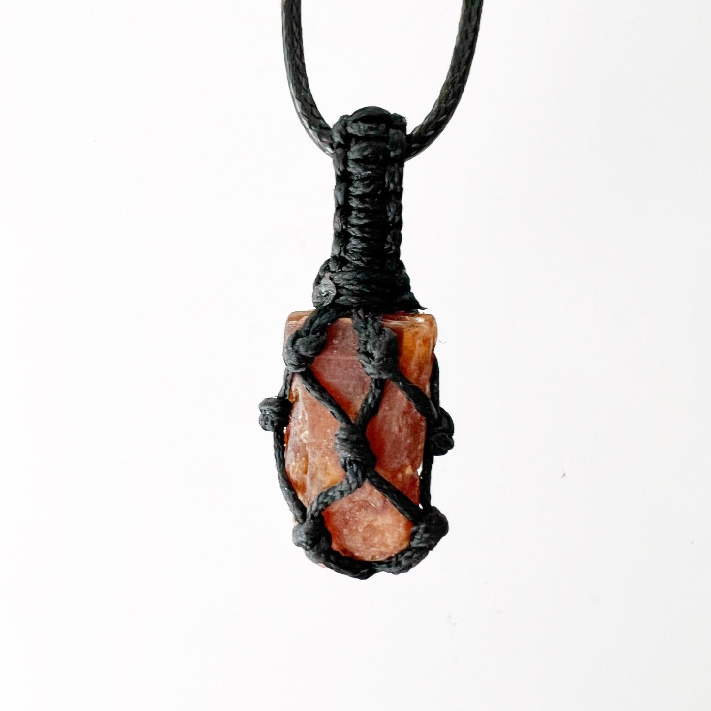 A Rough Orange Kyanite Crystal Thread Cage Pendant wrapped in an intricate black macrame netting is displayed against a white background. The detailed craftsmanship of the macrame cord securely encloses the irregularly shaped crystal, often associated with balancing the Sacral Chakra and enhancing creative thinking.