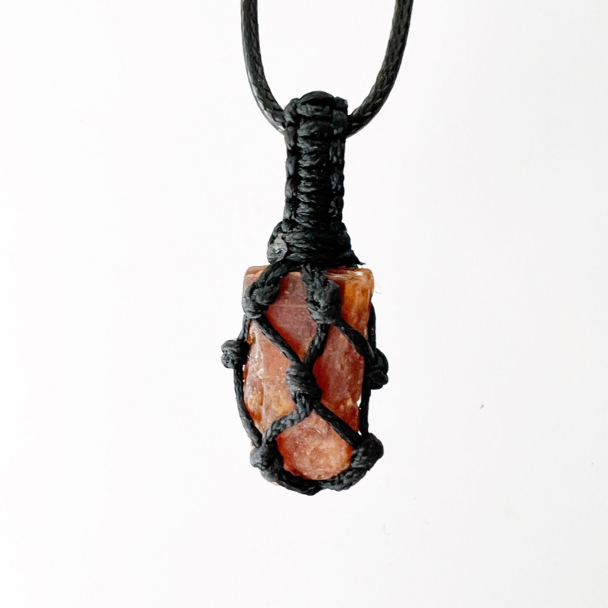 A Rough Orange Kyanite Crystal Thread Cage Pendant wrapped in an intricate black macrame netting is displayed against a white background. The detailed craftsmanship of the macrame cord securely encloses the irregularly shaped crystal, often associated with balancing the Sacral Chakra and enhancing creative thinking.