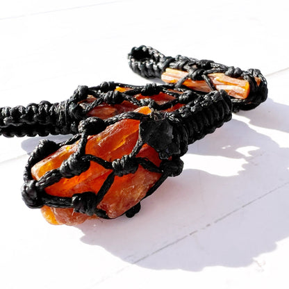 A Rough Orange Kyanite Crystal Thread Cage Pendant wrapped in an intricate black macrame netting is displayed against a white background. The detailed craftsmanship of the macrame cord securely encloses the irregularly shaped crystal, often associated with balancing the Sacral Chakra and enhancing creative thinking.