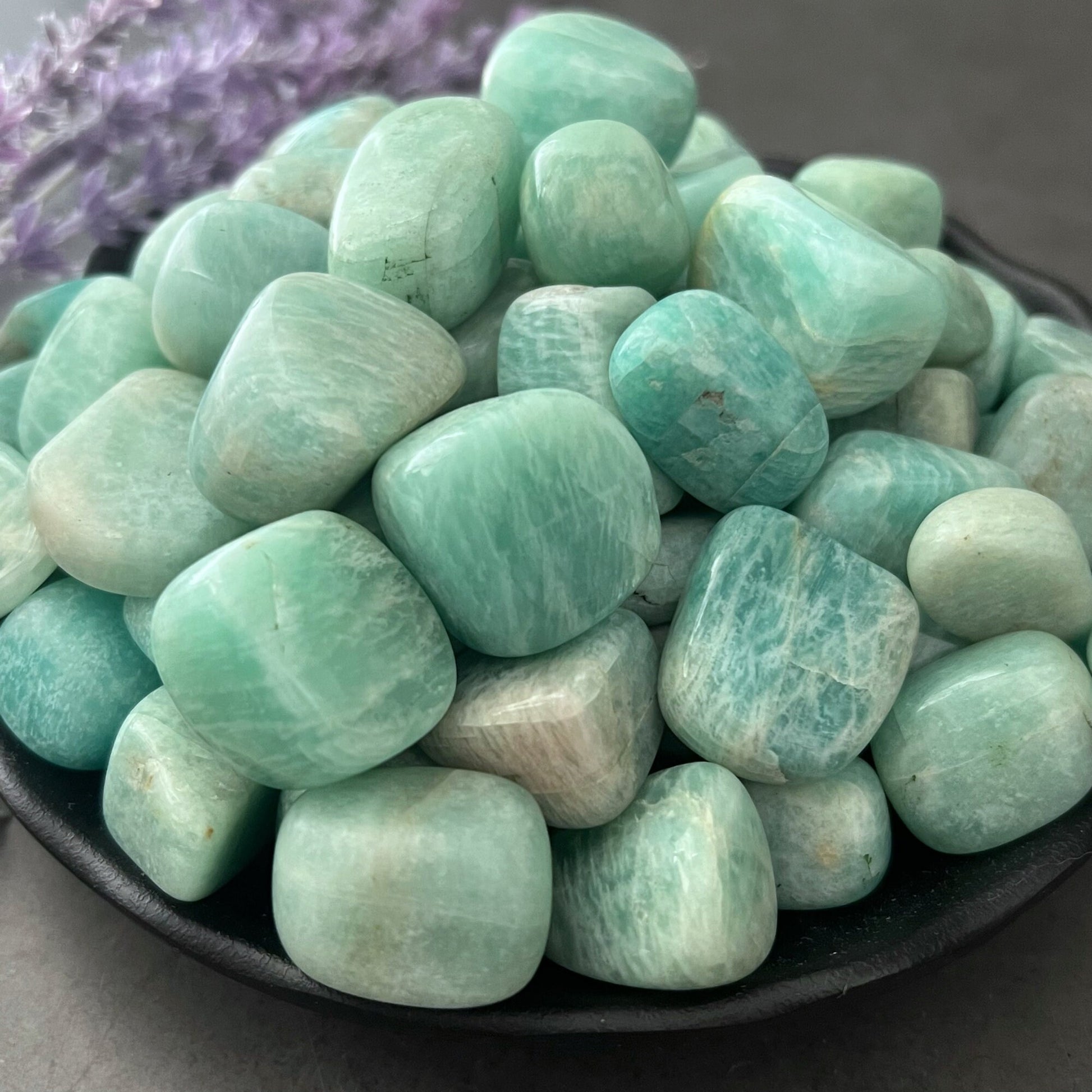 A black bowl filled with polished Amazonite Tumbled Stones sits on a smooth gray surface. To the left of the bowl, sprigs of purple lavender are visible, imparting calming energy and adding a touch of color to the composition.