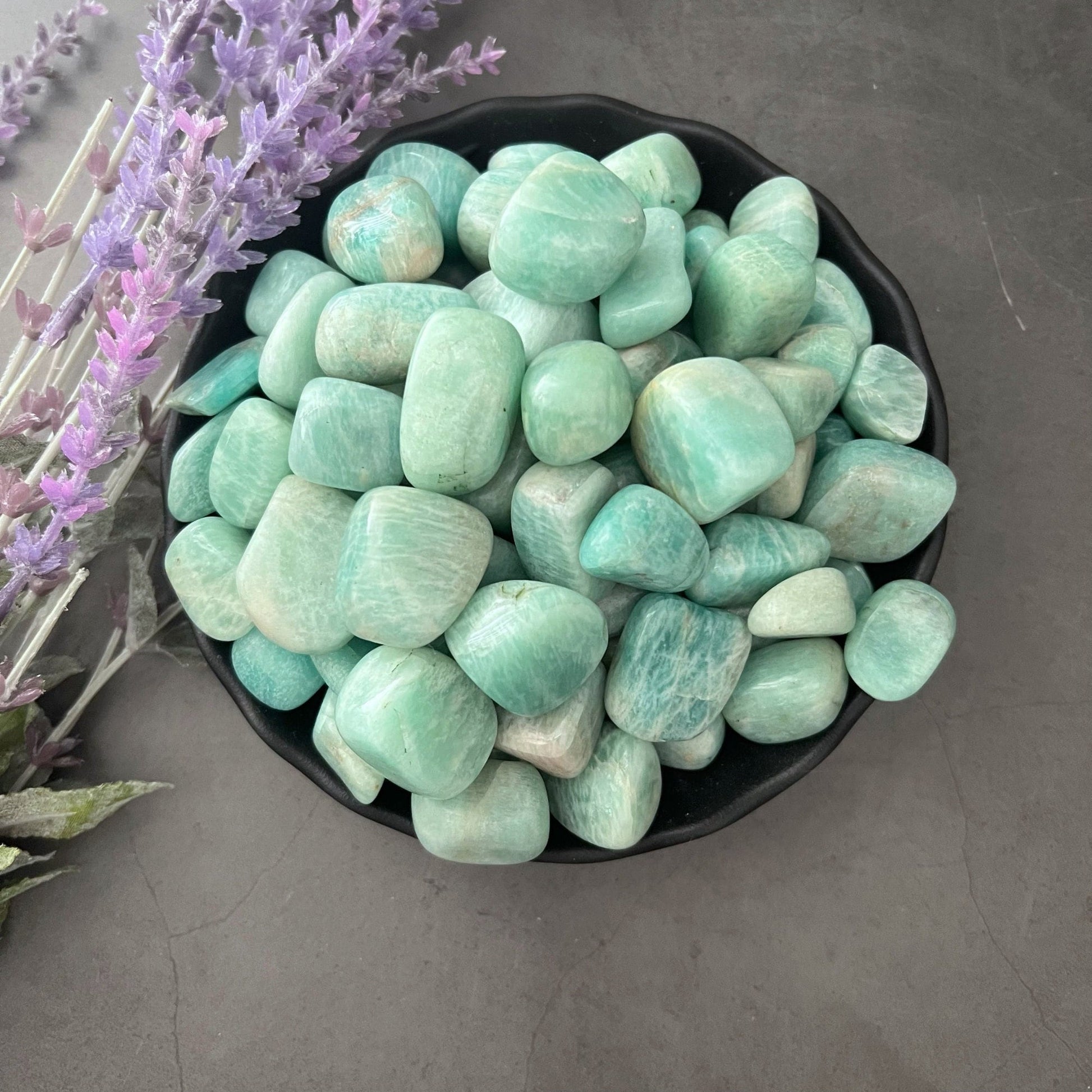 A black bowl filled with polished Amazonite Tumbled Stones sits on a smooth gray surface. To the left of the bowl, sprigs of purple lavender are visible, imparting calming energy and adding a touch of color to the composition.