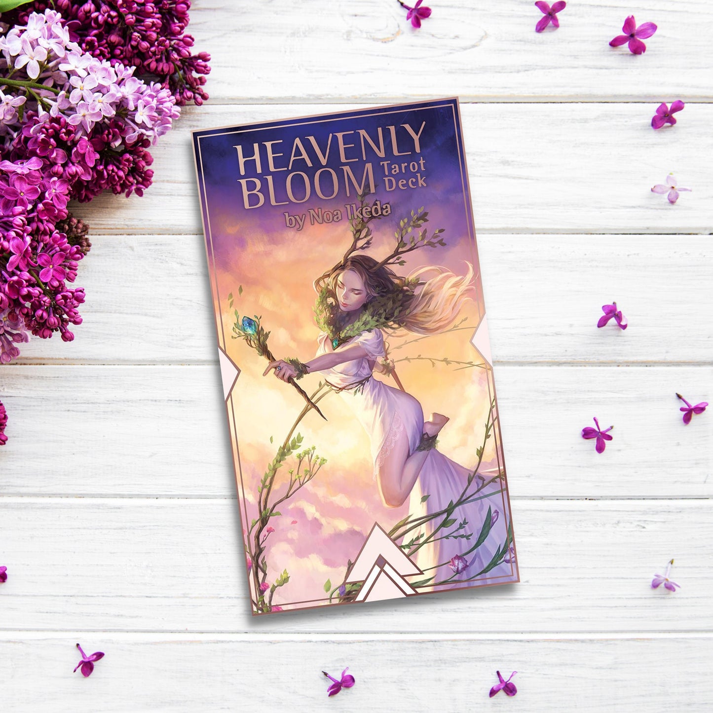 A boxed set of "Heavenly Bloom Tarot" by Noa Ikeda is placed on a white wooden surface surrounded by scattered purple flowers and lilac blossoms. The box, adorned with floral elements, features an illustration of a woman with flowing hair holding a branch, set against a sunset sky.