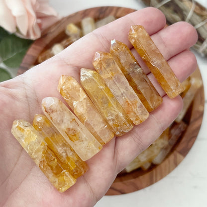 A wooden bowl filled with 1-2" Golden Healer Quartz Points in shades of yellow and clear sits on a white surface. To the left, there is a pink flower with leaves, and at the top right, an energy generator sage bundle wrapped with white string.