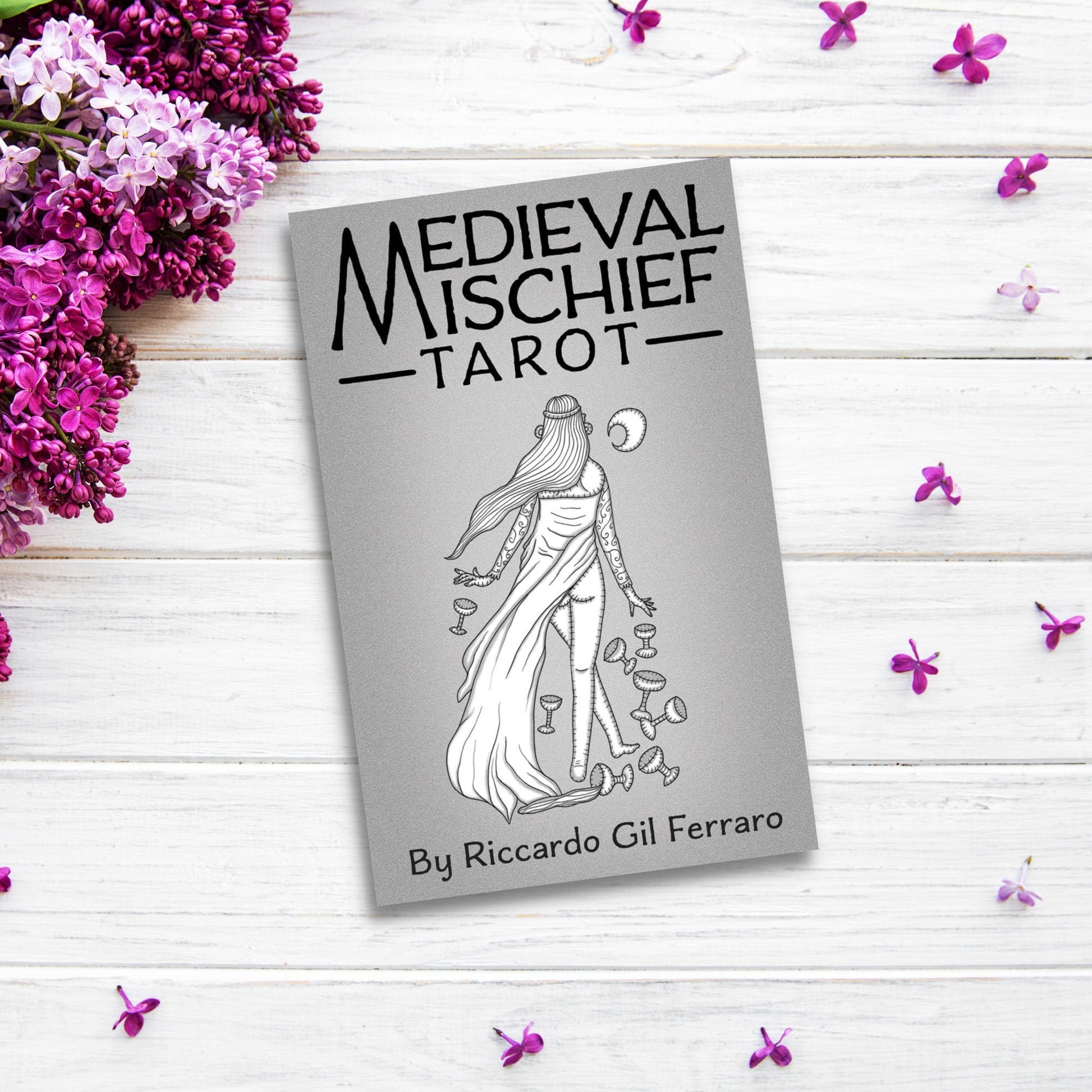 The image shows a "Medieval Mischief Tarot" by Riccardo Gil Ferraro placed on a white wooden surface. The cover features an illustration of a woman in a flowing dress surrounded by several cups and a crescent moon, with purple flowers around the card, embodying the allure of esoteric tarot decks.