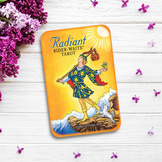 A Tarot card deck titled "Radiant Rider-Waite® in a Tin" sits on a white wooden surface surrounded by purple and white flowers. The cover illustration features a colorful figure standing on a cliff holding a flower, with a small dog beside them and a bright sun overhead. This edition comes in a charming keepsake tin.
