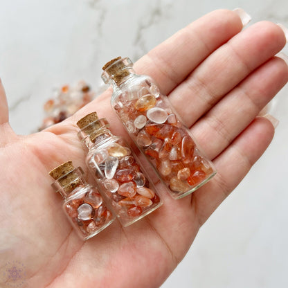 A wooden bowl filled with various tumbled and polished stones sits on a marble surface. Surrounding the bowl are three small glass bottles containing Hematoid Quartz, a bundle of red sage, and a plastic bag with Fire Quartz Crystal Chips.