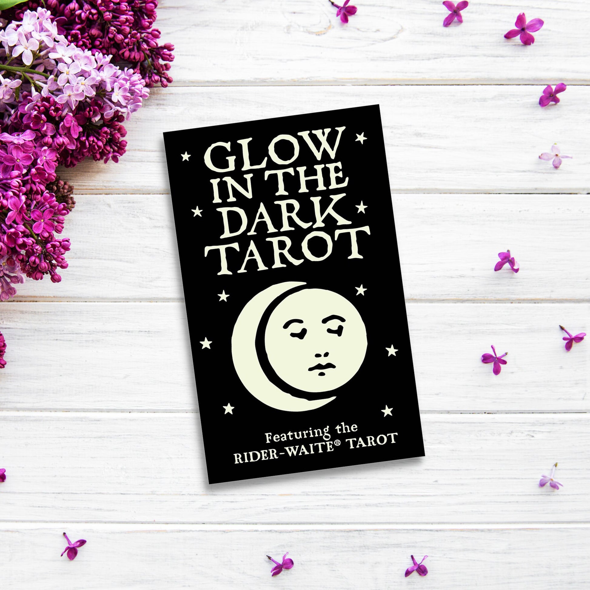 A tarot card box labeled "Glow in the Dark Tarot" is displayed on a white wooden surface. The black box adorned with stars and a moon design shines under low light. Purple flowers border the left side, with scattered petals on the right, giving a mystical feel.