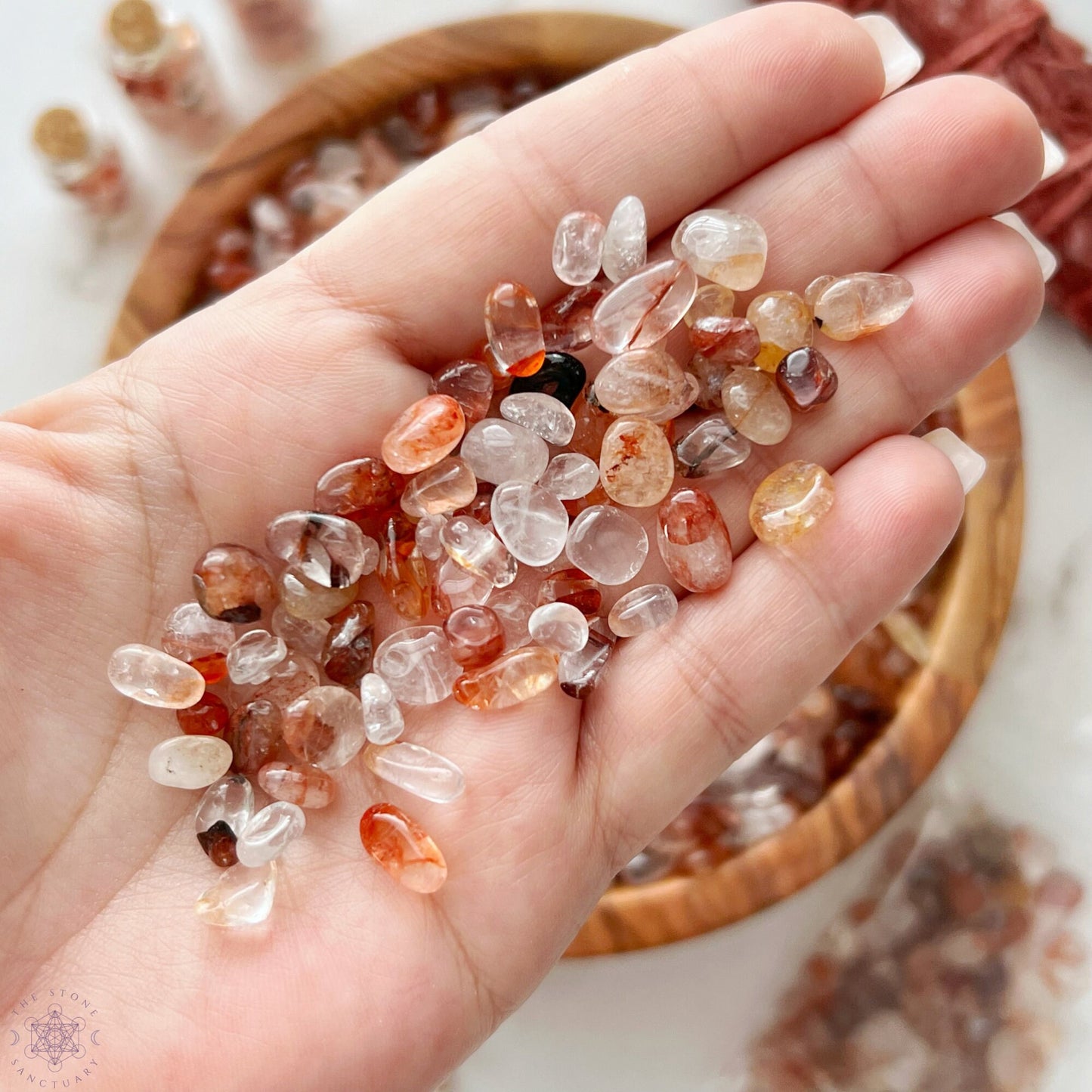 A wooden bowl filled with various tumbled and polished stones sits on a marble surface. Surrounding the bowl are three small glass bottles containing Hematoid Quartz, a bundle of red sage, and a plastic bag with Fire Quartz Crystal Chips.
