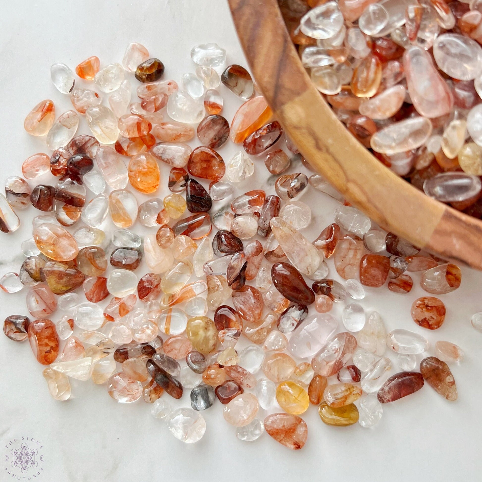 A wooden bowl filled with various tumbled and polished stones sits on a marble surface. Surrounding the bowl are three small glass bottles containing Hematoid Quartz, a bundle of red sage, and a plastic bag with Fire Quartz Crystal Chips.