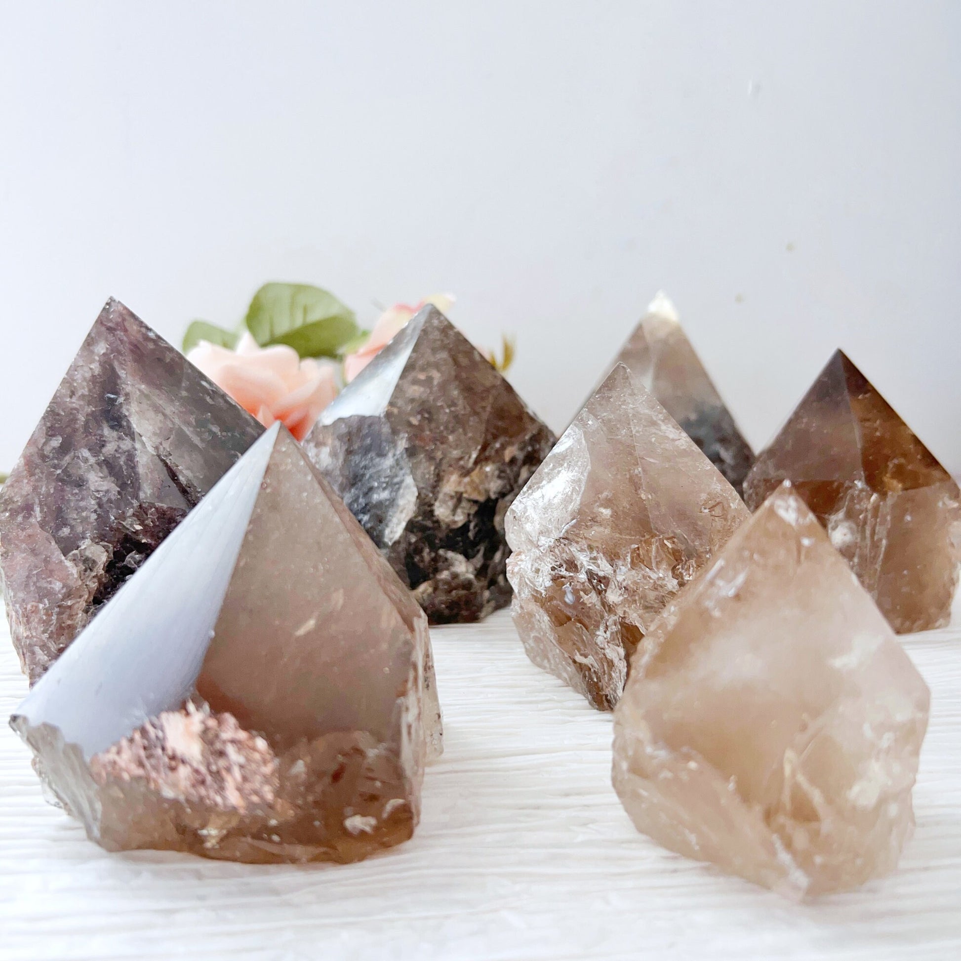 A close-up image features several 2-4" Smoky Quartz Top Polished Points, arranged meticulously in a crystal grid on a light-colored surface. The crystals vary in size and color intensity, with some exhibiting clear and reflective surfaces. Blurred flowers are visible in the background.