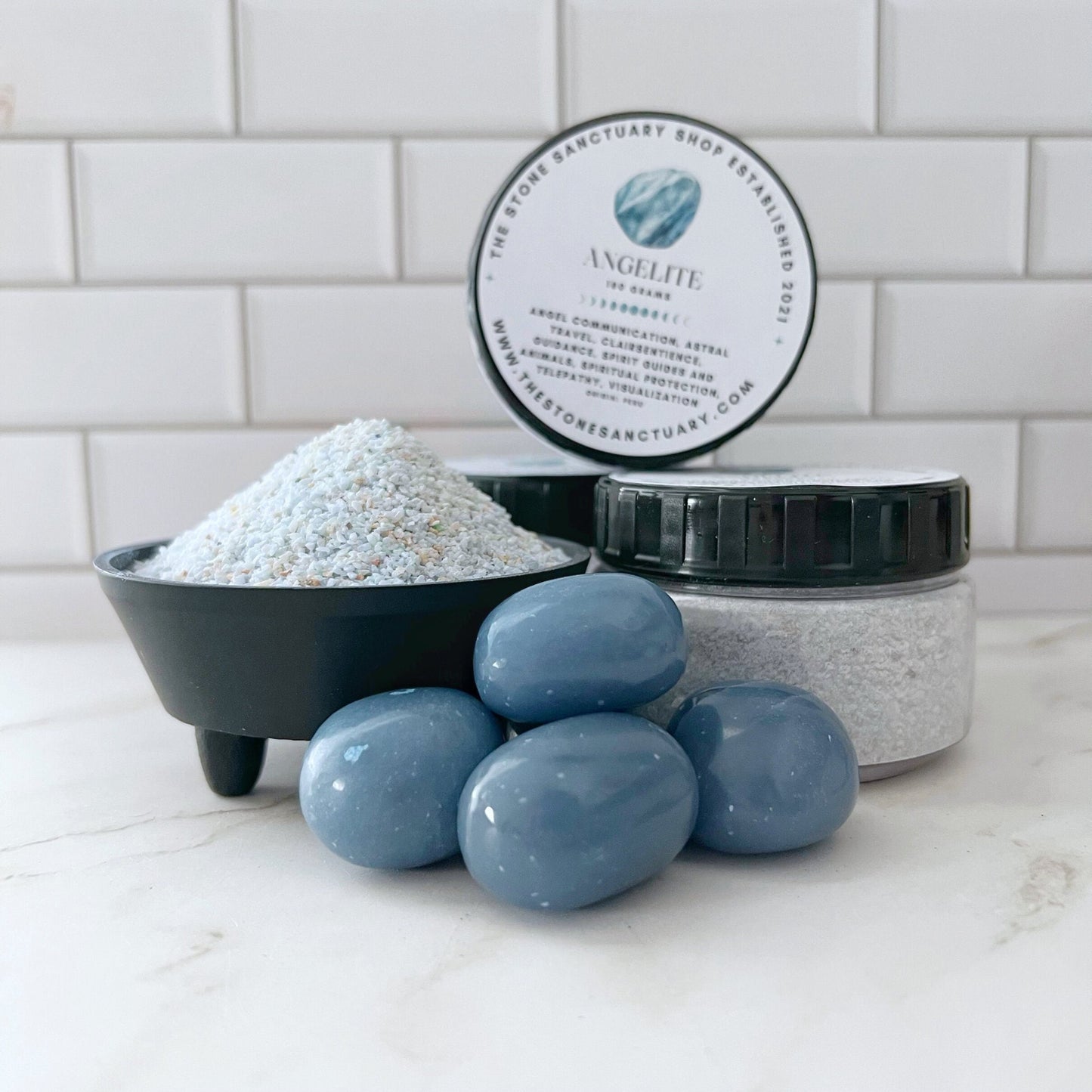 A jar of Angelite Gemstone Sand is displayed behind a small black bowl filled with the scrub and next to a pile of four smooth, blue gemstones that symbolize spiritual connection. The scene is set against a white tiled backsplash and a white countertop.