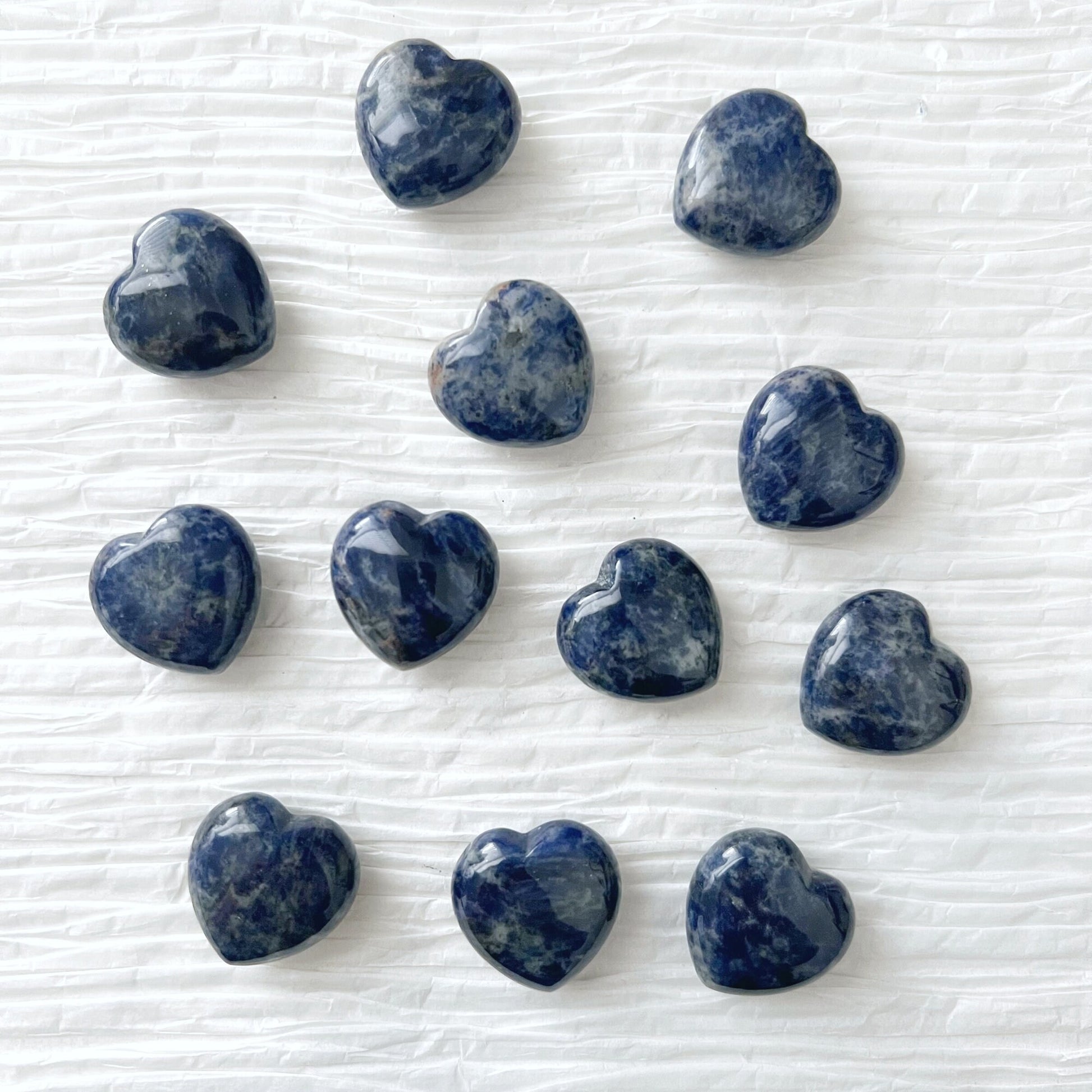 A round white plate holds five polished, heart-shaped stones, including a radiant 1" Mini Sodalite Heart. In the top left corner, a gold-rimmed dish is partially visible, also containing similarly shaped treasures. The background features white textured surfaces and a piece of paper with cursive writing, fostering inner peace.