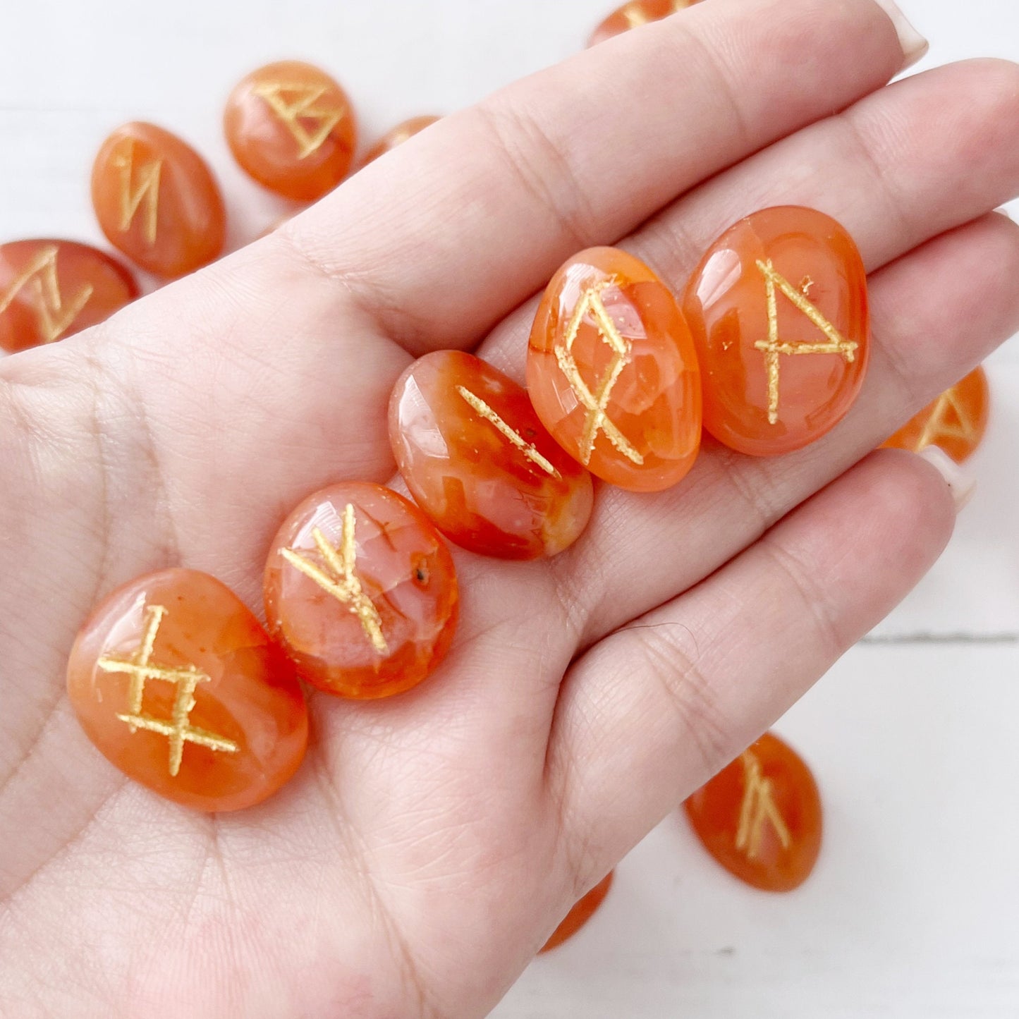 A display featuring an arrangement of orange Carnelian Elder Futhark Runes stones etched with gold symbols, a bundle of dried sage tied with string, a ceramic white skull, and a small pink succulent plant in a white pot. The items are set on a white surface, creating an enchanting scene for divination practices.
