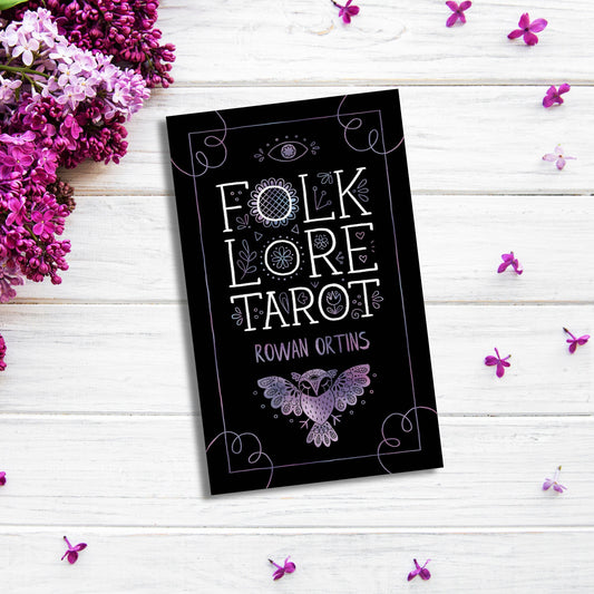 A black tarot card deck box titled "Folklore Tarot" by Rowan Ortins lies on a white wooden surface surrounded by purple and lilac flowers. The box features decorative white and purple designs, including an eye, floral patterns, and a bird. It comes with an illustrated guidebook exploring mythological traditions.