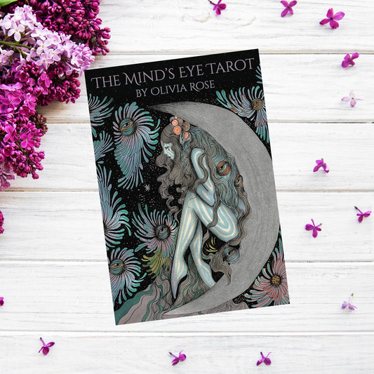 A tarot card deck box titled *The Mind's Eye Tarot* by Olivia Rose lies on a white wooden surface, surrounded by scattered purple flower petals and blooming lilac flowers. Alongside the box, which features an intricate illustration of a woman embracing a crescent moon, is a 172-page guidebook.
